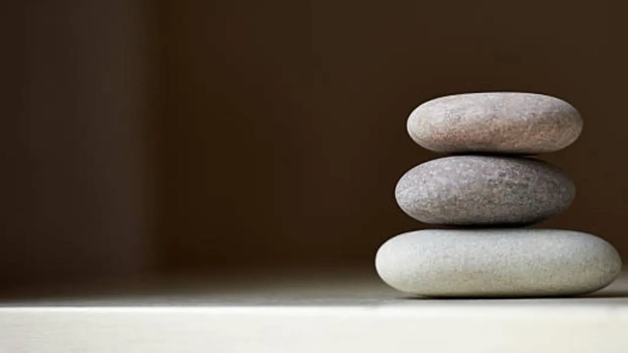 Three smooth, rounded stones are stacked on top of each other on a light surface, with a plain brown background and empty space to the left.