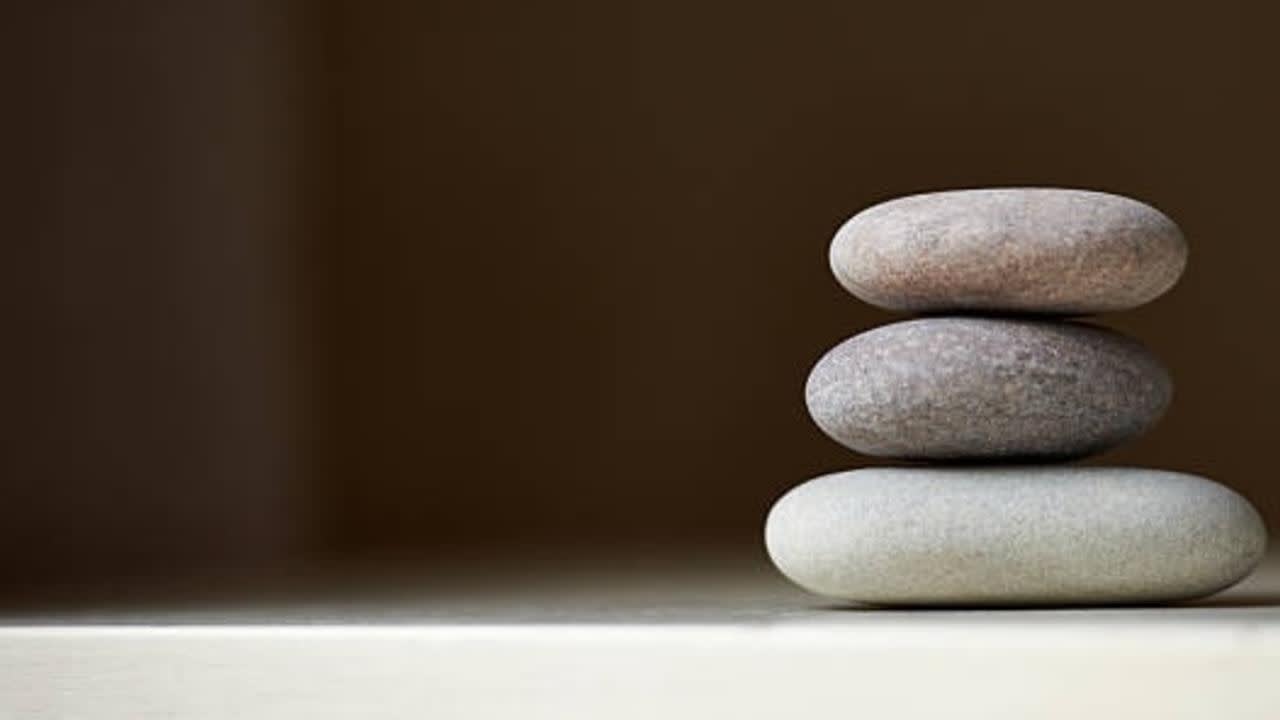 Three smooth, rounded stones are stacked on top of each other on a light surface, with a plain brown background and empty space to the left.