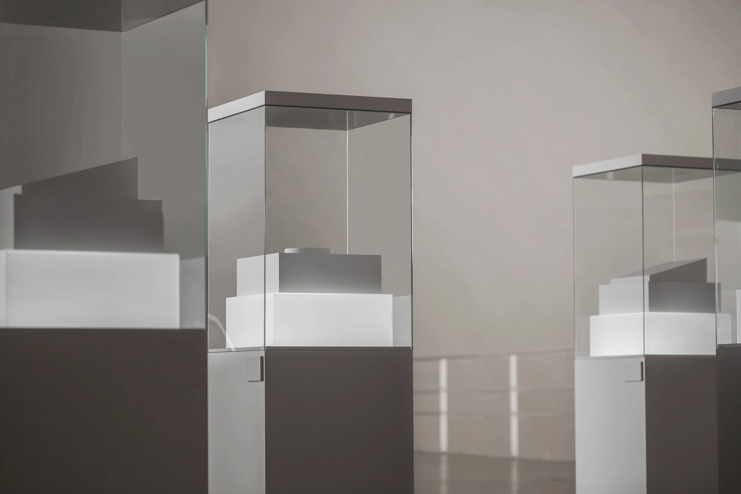 Glass display cases with minimal white geometric objects inside are arranged in a well-lit, modern exhibition space with a neutral background.