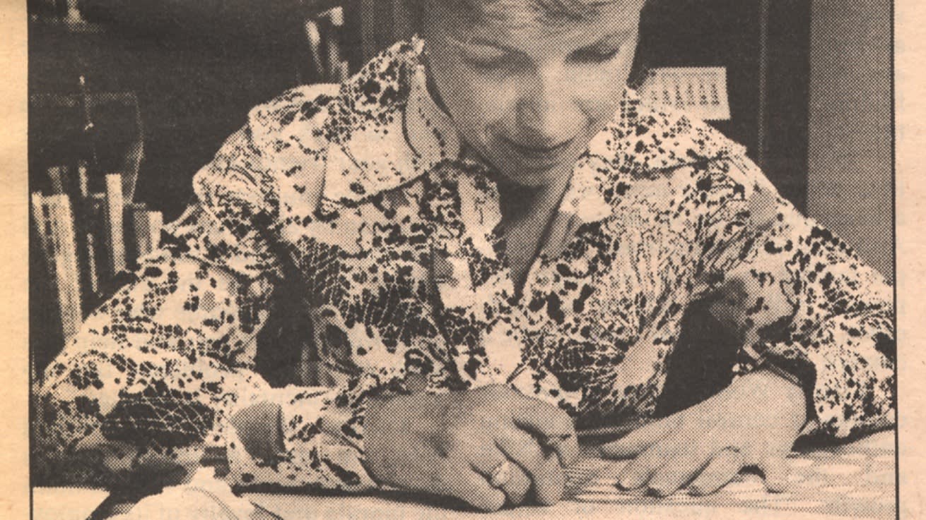 A woman wearing a patterned blouse is seated at a desk, writing or drawing on a piece of paper with a pen. Books and office items are visible in the background. The image has a vintage, sepia tone.