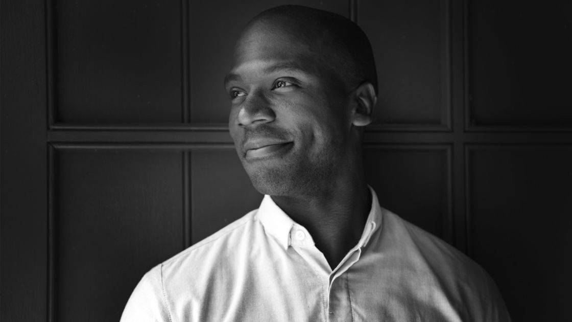 Black and white photo of a man in a light button-up shirt, looking to the side and smiling softly, standing in front of a paneled wall.