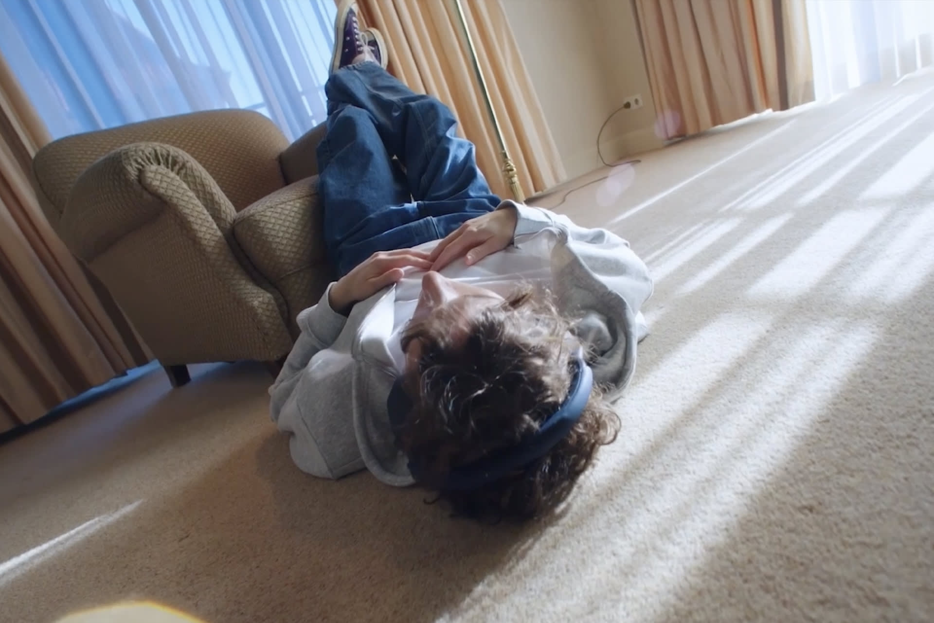A person wearing jeans, a gray sweater, and a blue headband lies on their back on a carpeted floor with legs propped up on a chair, in a sunlit room with curtains and large windows.