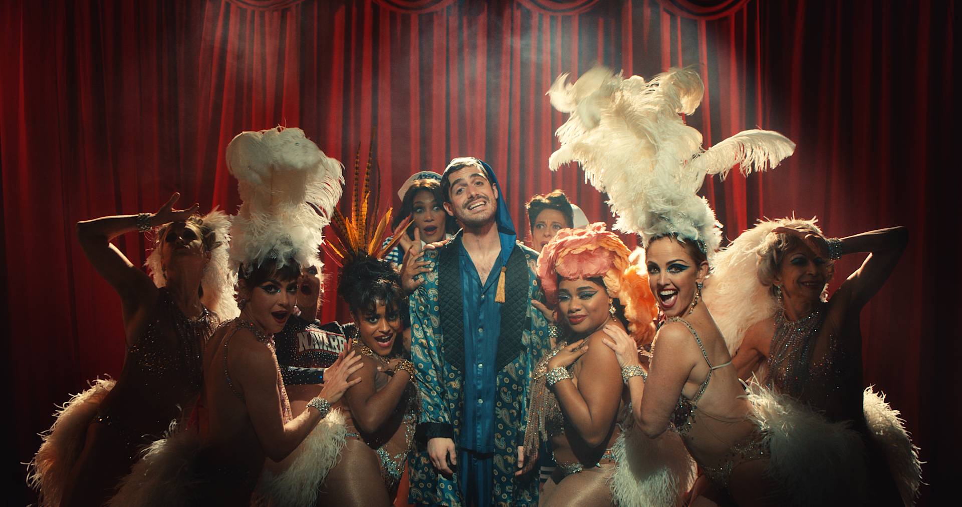 A smiling man in a blue robe stands center stage, surrounded by showgirls in glamorous costumes with feathered headdresses and fans, in front of a red curtain under dramatic theater lighting.