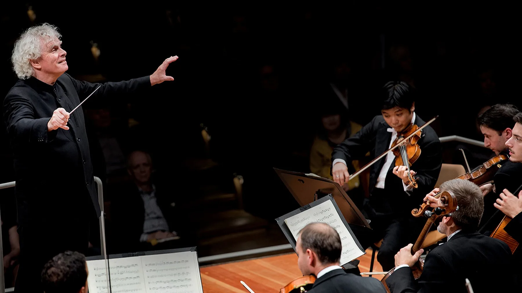 A conductor energetically leads an orchestra, with several musicians playing violins and cellos on stage, illuminated under bright lights against a dark background.