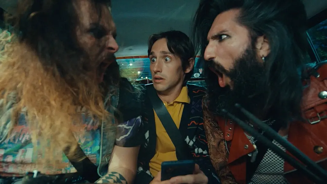 Three men sit in a car; the two on the sides, dressed in bold, rock-style outfits, are angrily shouting, while the man in the middle, wearing a yellow shirt, looks surprised and uncomfortable, holding a phone.