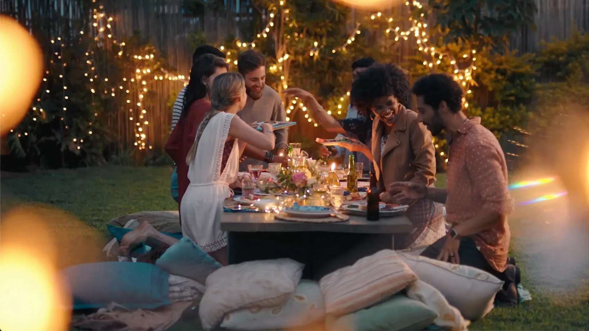 A group of friends share a meal at a low outdoor table surrounded by string lights and cushions in a backyard, creating a festive and cozy evening atmosphere.