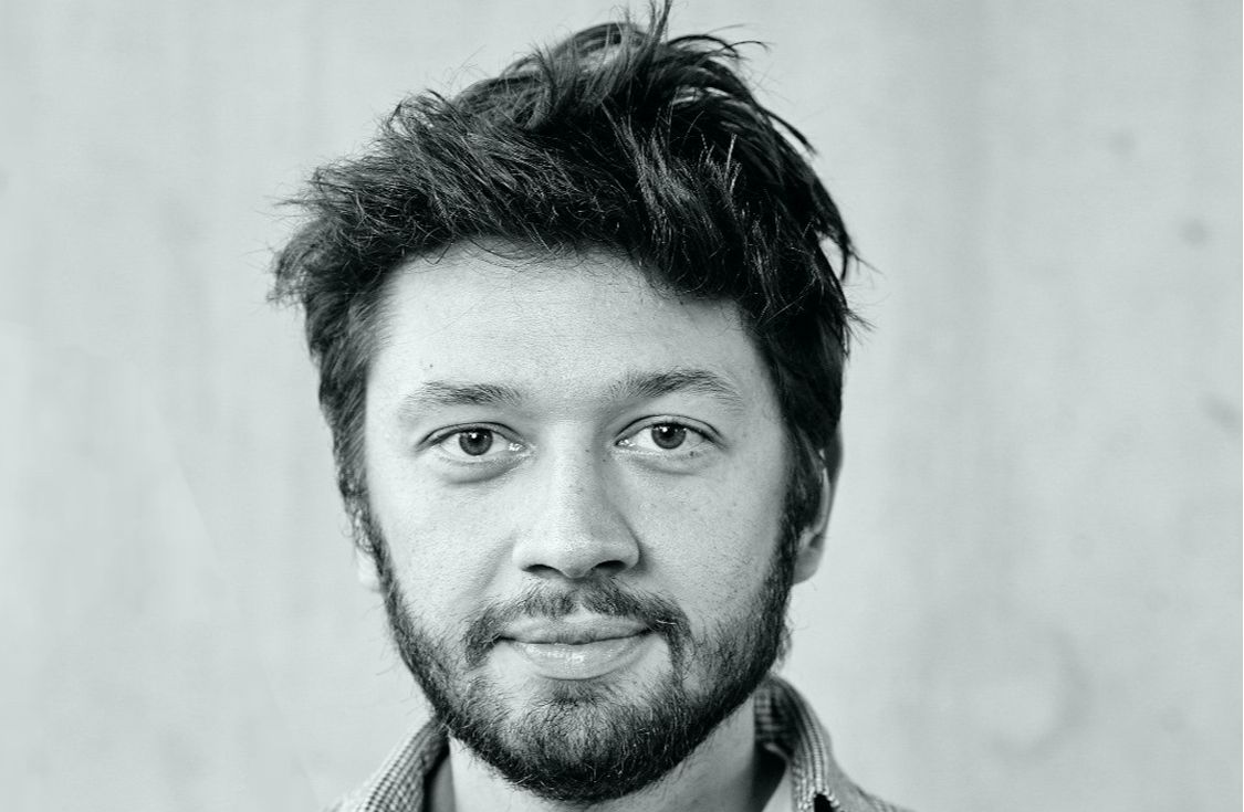 A young man with dark messy hair and a beard looks directly at the camera with a neutral expression. He is wearing a light-colored, collared shirt and the background is plain and out of focus.