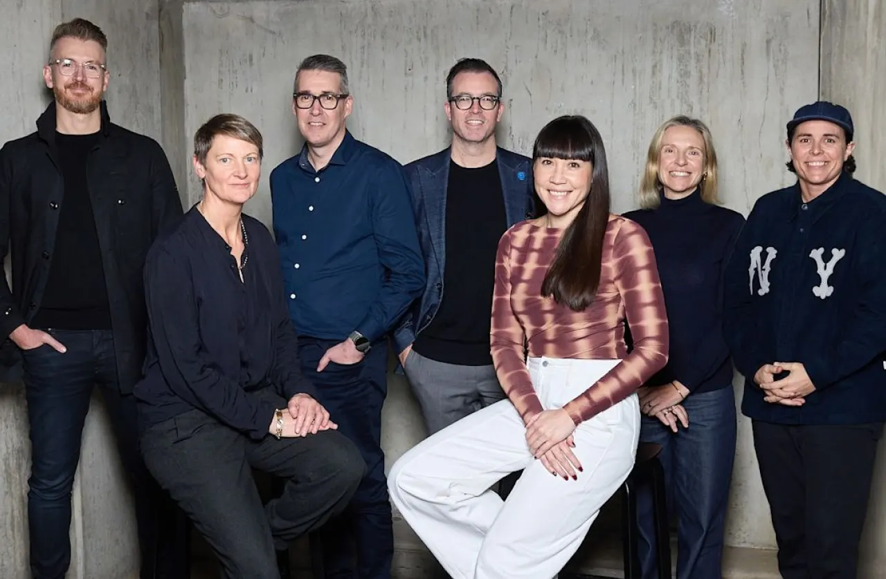 Seven people pose together indoors against a plain concrete wall. Four stand in the back and three sit in front. They are casually dressed and smiling, facing the camera.