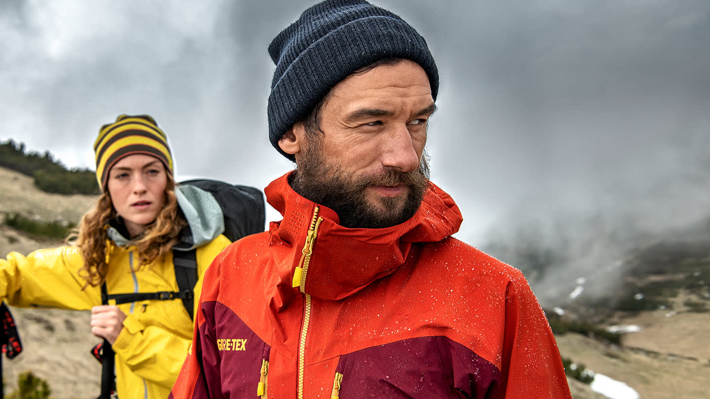 A bearded man in a red jacket and navy beanie stands outdoors in rainy, mountainous terrain, with a woman in a yellow jacket and striped hat behind him, both looking into the distance.