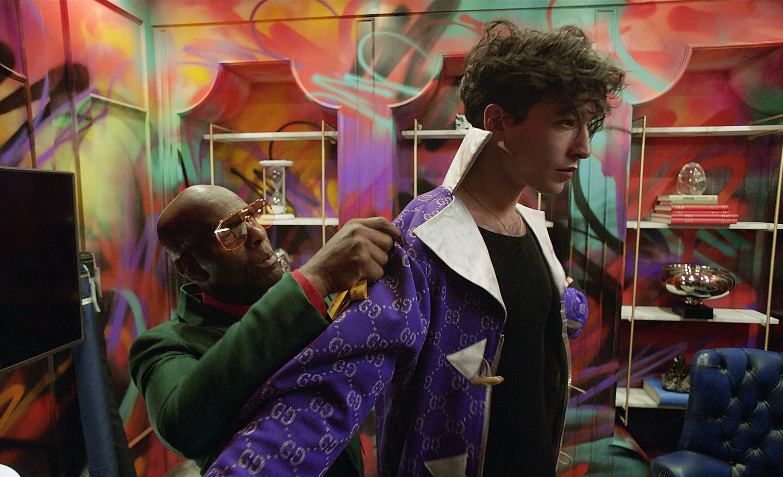 A man in sunglasses adjusts the sleeve of a purple Gucci jacket worn by another man standing in front of him, inside a colorful, graffiti-covered room with shelves and books in the background.