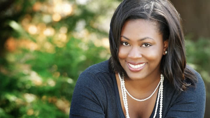 A woman with shoulder-length dark hair smiles at the camera. She is wearing a dark top and a pearl necklace, with greenery and soft sunlight in the blurred background.