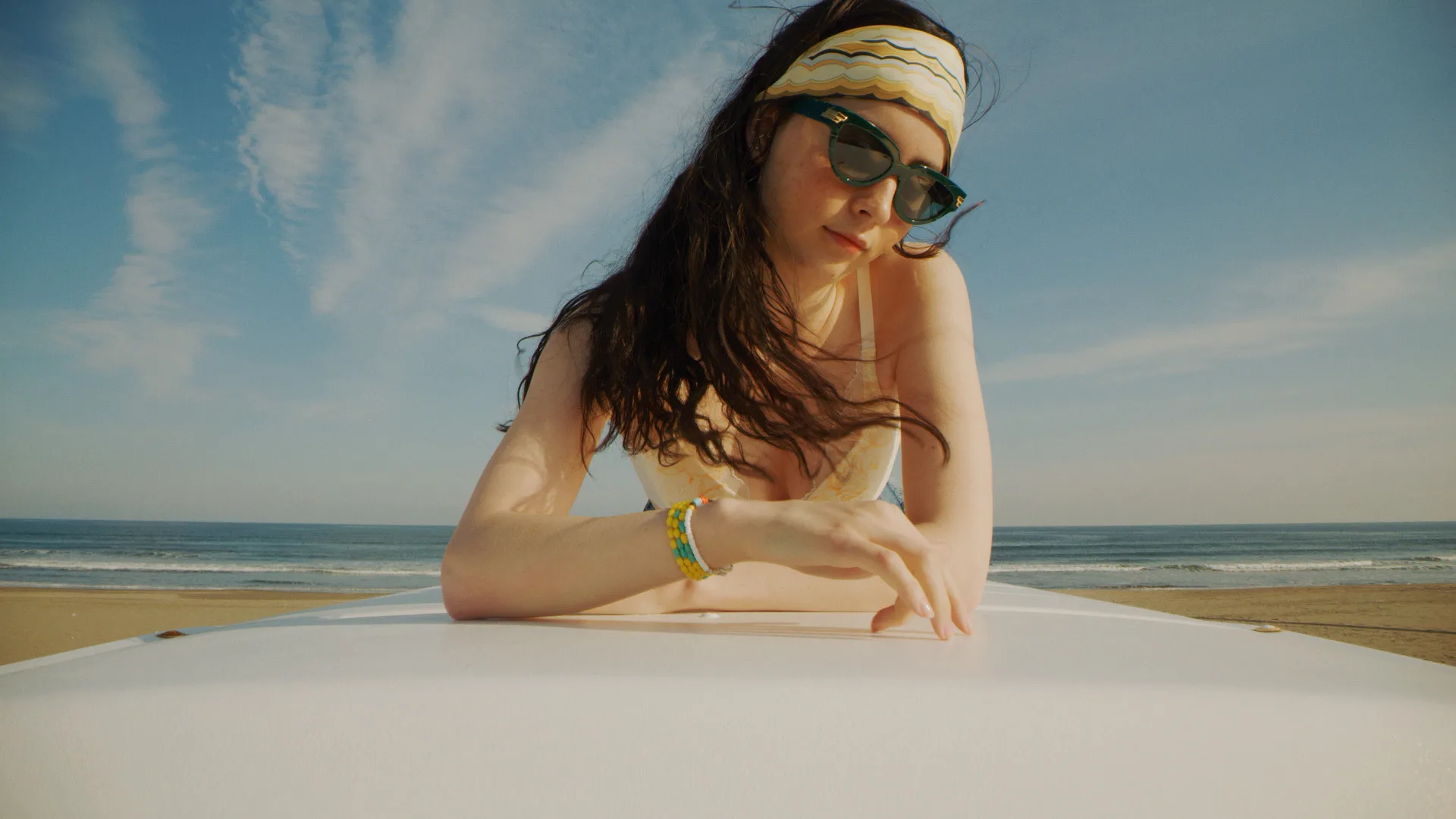 A woman wearing sunglasses and a headband leans on a white surface at the beach, with the ocean and blue sky in the background. She has long dark hair and is wearing colorful bracelets.