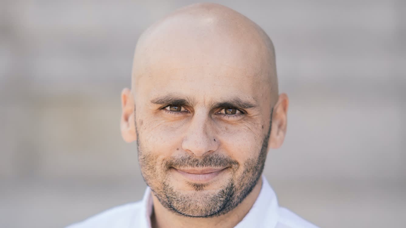A bald man with a trimmed beard and mustache, wearing a white shirt, smiling gently at the camera with a blurred gray background.