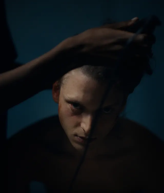 A person with a serious expression looks up as another hand shaves their head with electric clippers, set against a dark, moody background.