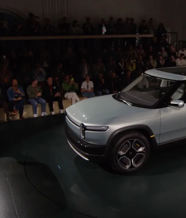 A light blue Rivian electric truck is showcased on a circular platform indoors, with a large seated audience watching in the background.