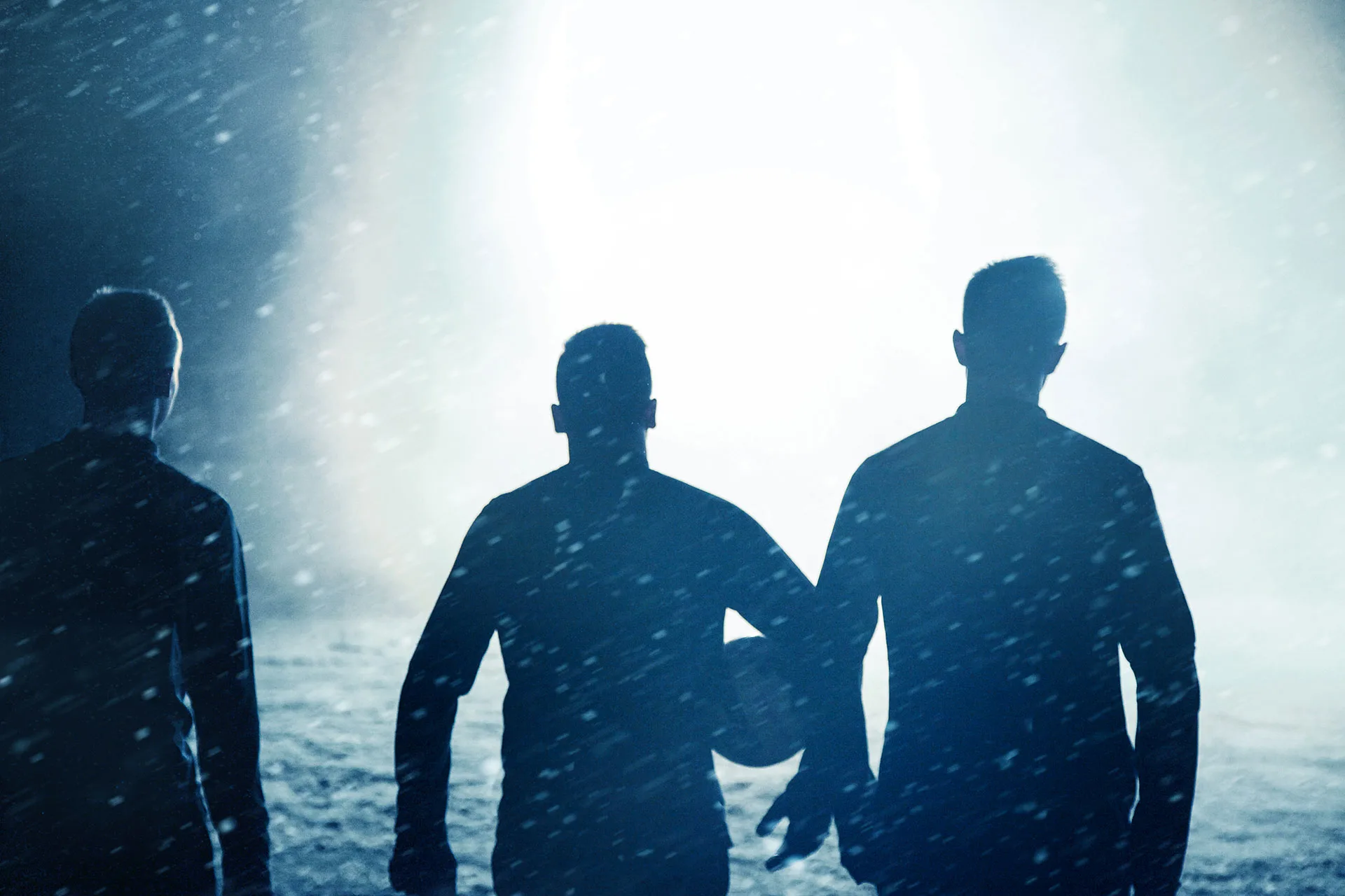 Three people are seen from behind, standing close together in a snowy environment at night, illuminated by bright light in the background. One person in the center is holding a ball.