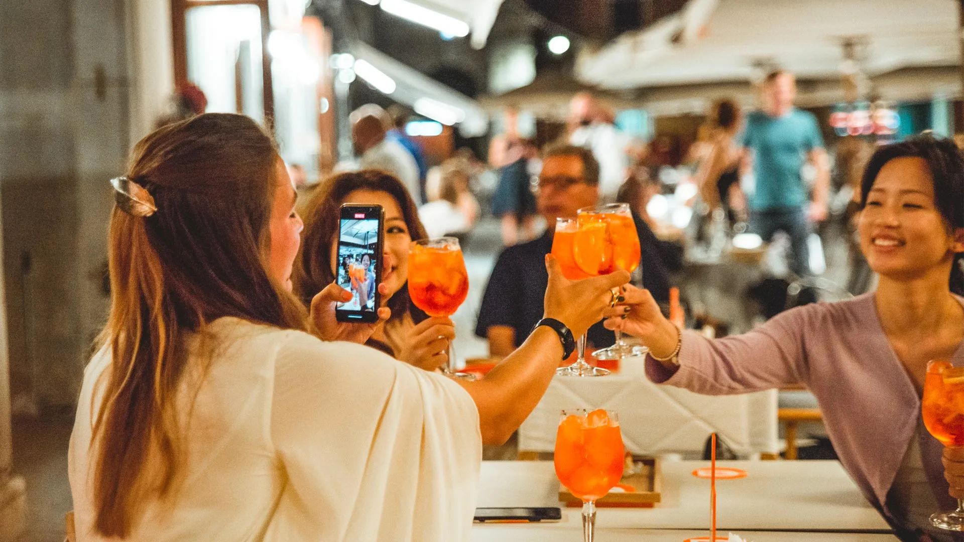 Four women sit at an outdoor restaurant, smiling and raising large glasses of orange cocktails for a toast. One woman holds up a smartphone to capture the moment. Other diners are visible in the background.