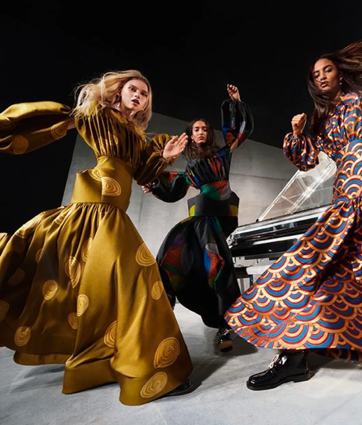 Three women in bold, patterned dresses pose dynamically in a modern setting with geometric walls and a piano, creating a sense of movement and energy.