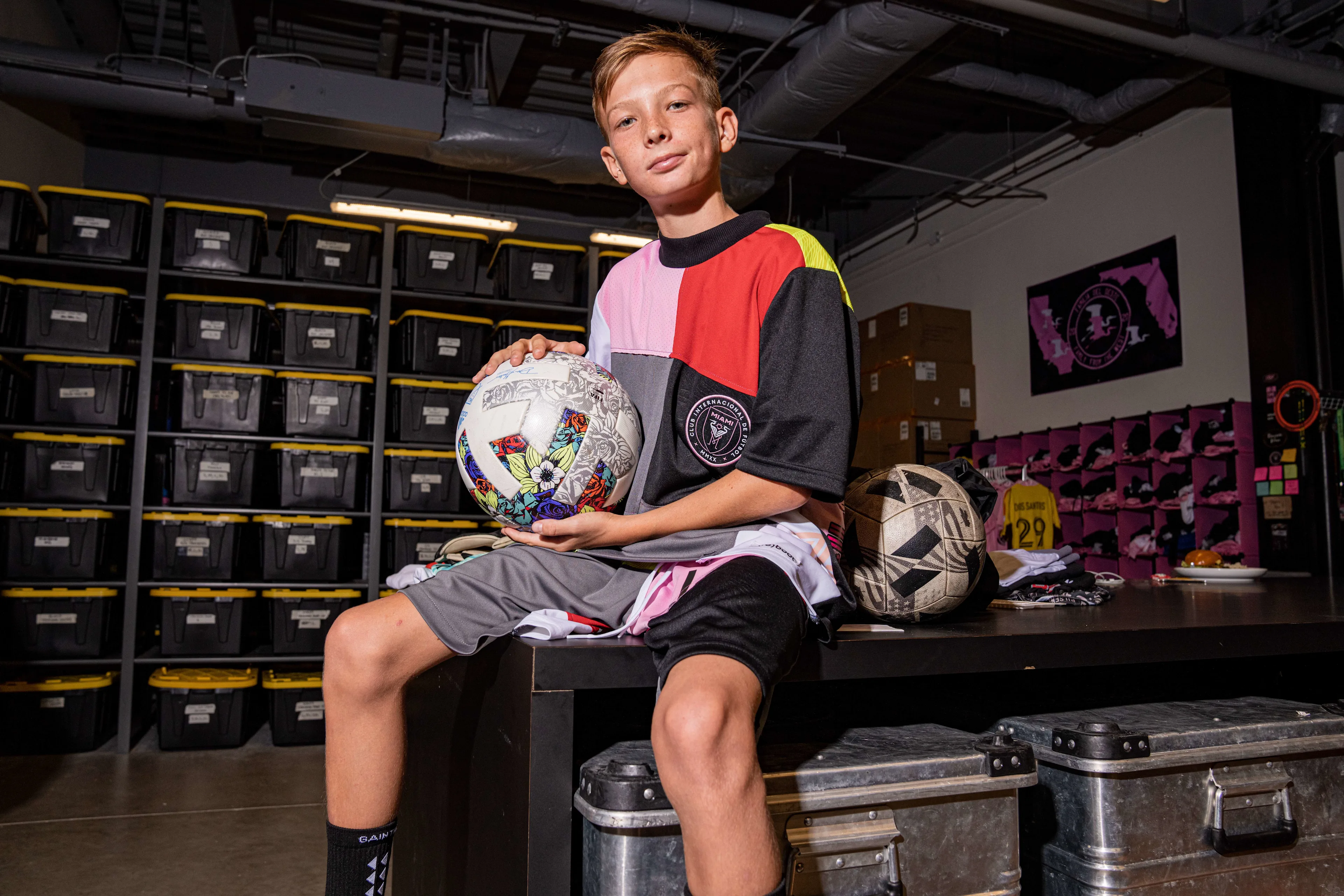 A boy sits on a table in a storage room, holding a decorated soccer ball. He wears a colorful shirt and black shorts. Behind him are stacked black storage bins and soccer equipment.