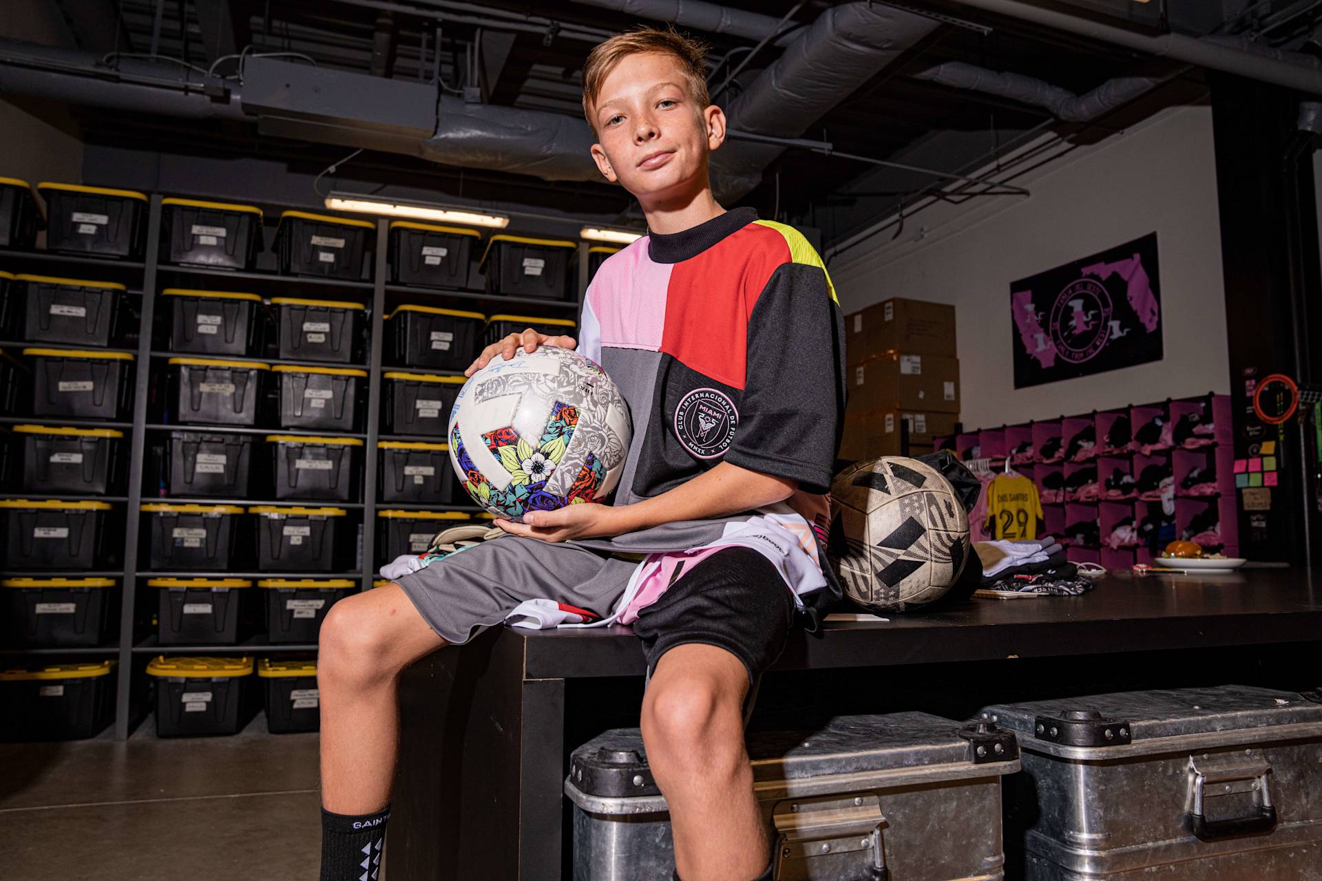 A boy sits on a table in a storage room, holding a decorated soccer ball. He wears a colorful shirt and black shorts. Behind him are stacked black storage bins and soccer equipment.