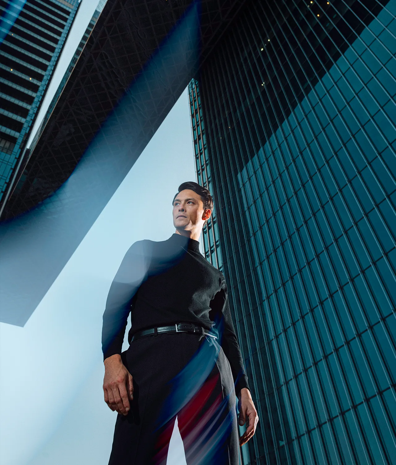 A man in a black turtleneck and trousers stands between tall modern glass buildings, looking confidently into the distance, with blue light streaks crossing in the foreground.