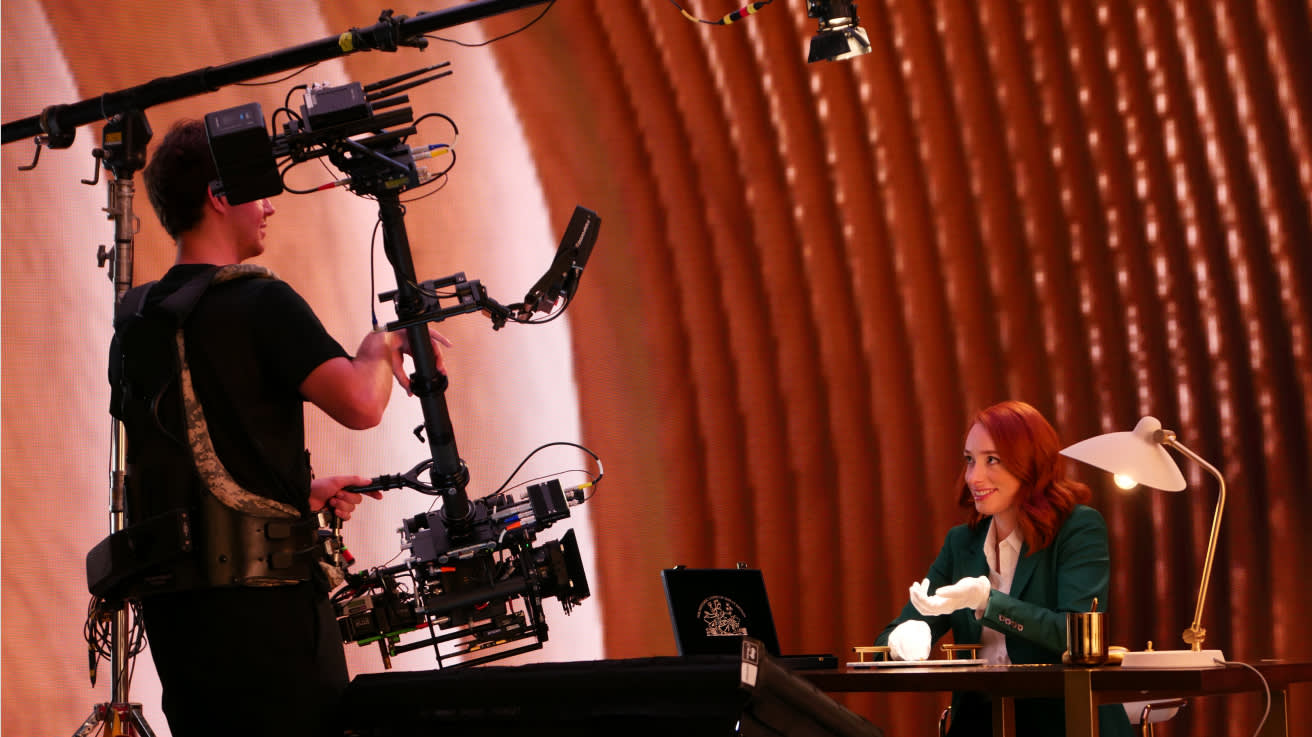 A camera operator films a woman with red hair, seated at a desk with a lamp and laptop, against a warmly lit, curved background on a set. The woman smiles and gestures with gloved hands.
