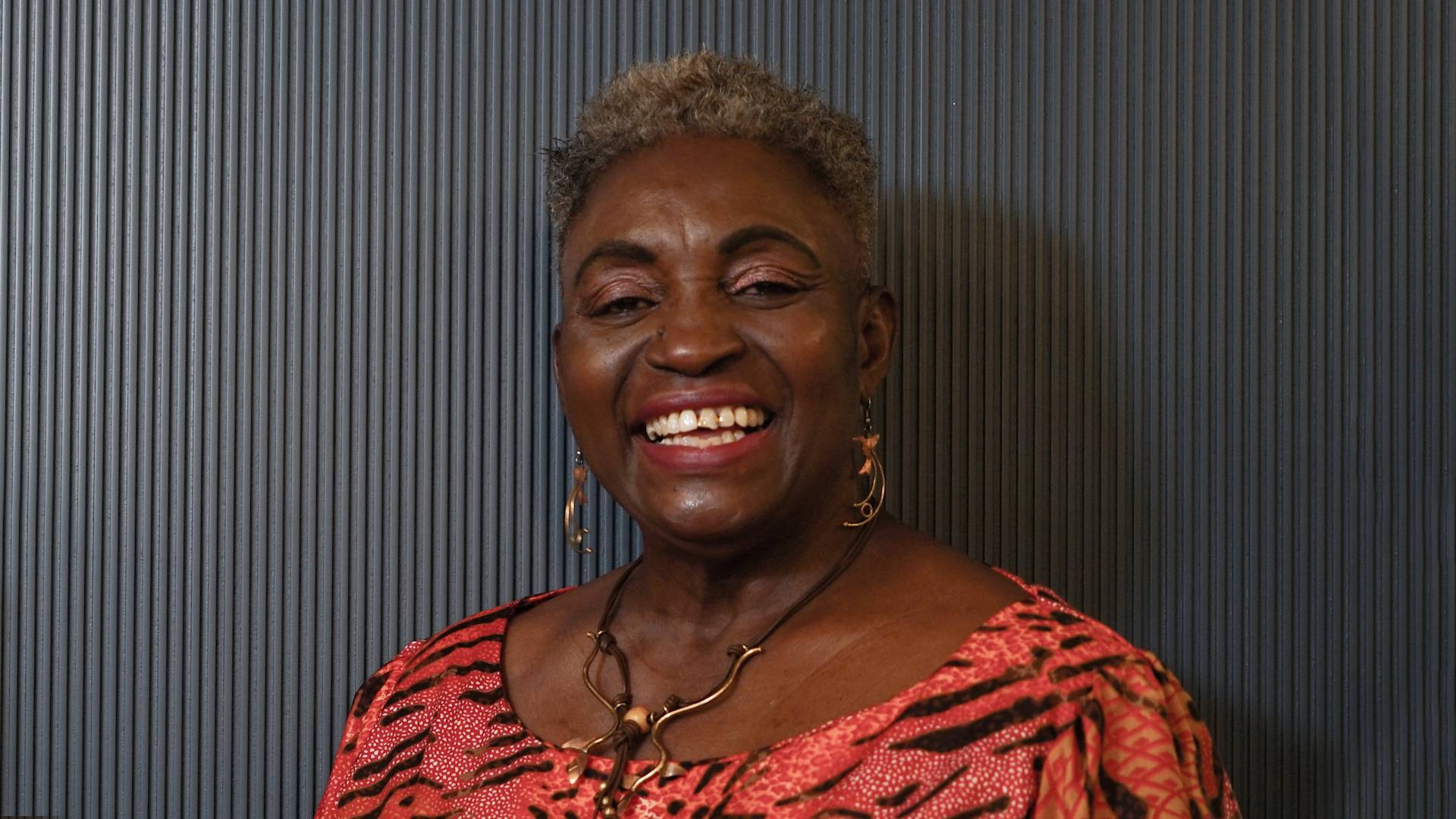 An older woman with short gray hair smiles warmly, wearing a patterned red and black top, gold earrings, and a gold necklace, standing in front of a dark, textured background.