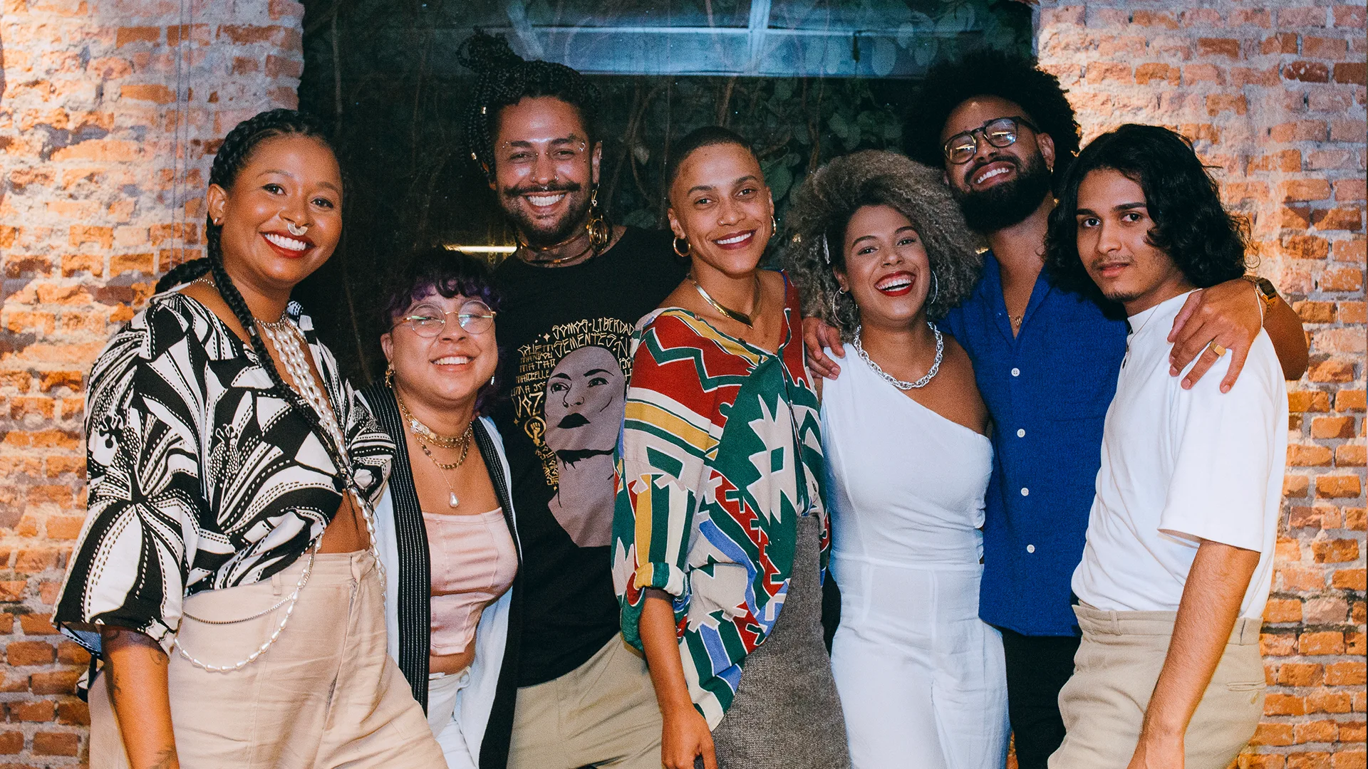 Seven people stand together smiling in front of a brick wall, with their arms around each other. They are dressed in colorful, stylish outfits and appear to be enjoying each others company.