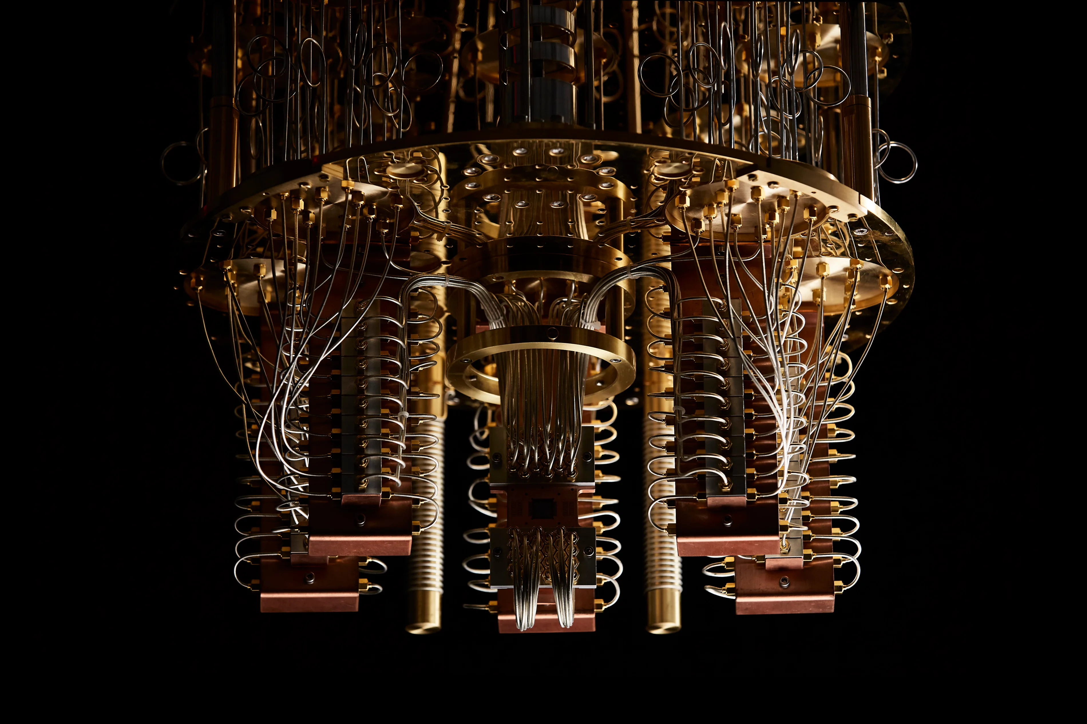 A close-up view of a quantum computer’s intricate hardware, featuring numerous gold and copper wires, coils, and components suspended in a complex, cylindrical structure against a black background.