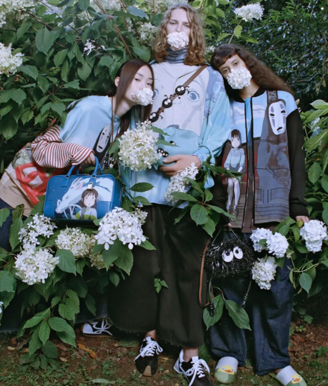 Three people stand amid leafy bushes and white flowers, with white blossoms covering their mouths. They wear colorful clothes and accessories featuring animated characters, blending with the surrounding lush greenery.