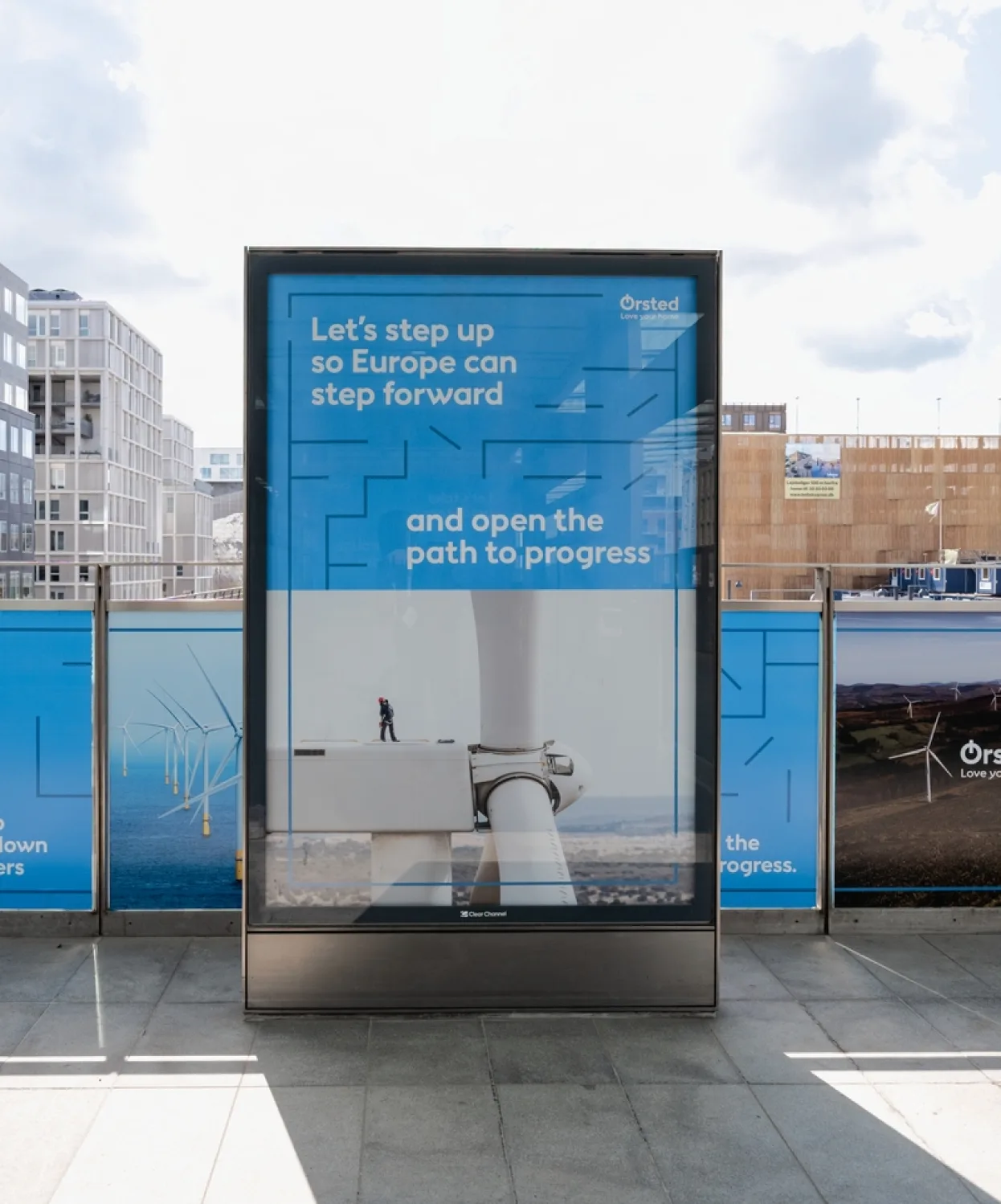 A digital billboard displays an Ørsted ad reading Lets step up so Europe can step forward and open the path to progress, with an image of a worker standing on a wind turbine. Urban buildings and blue sky are in the background.