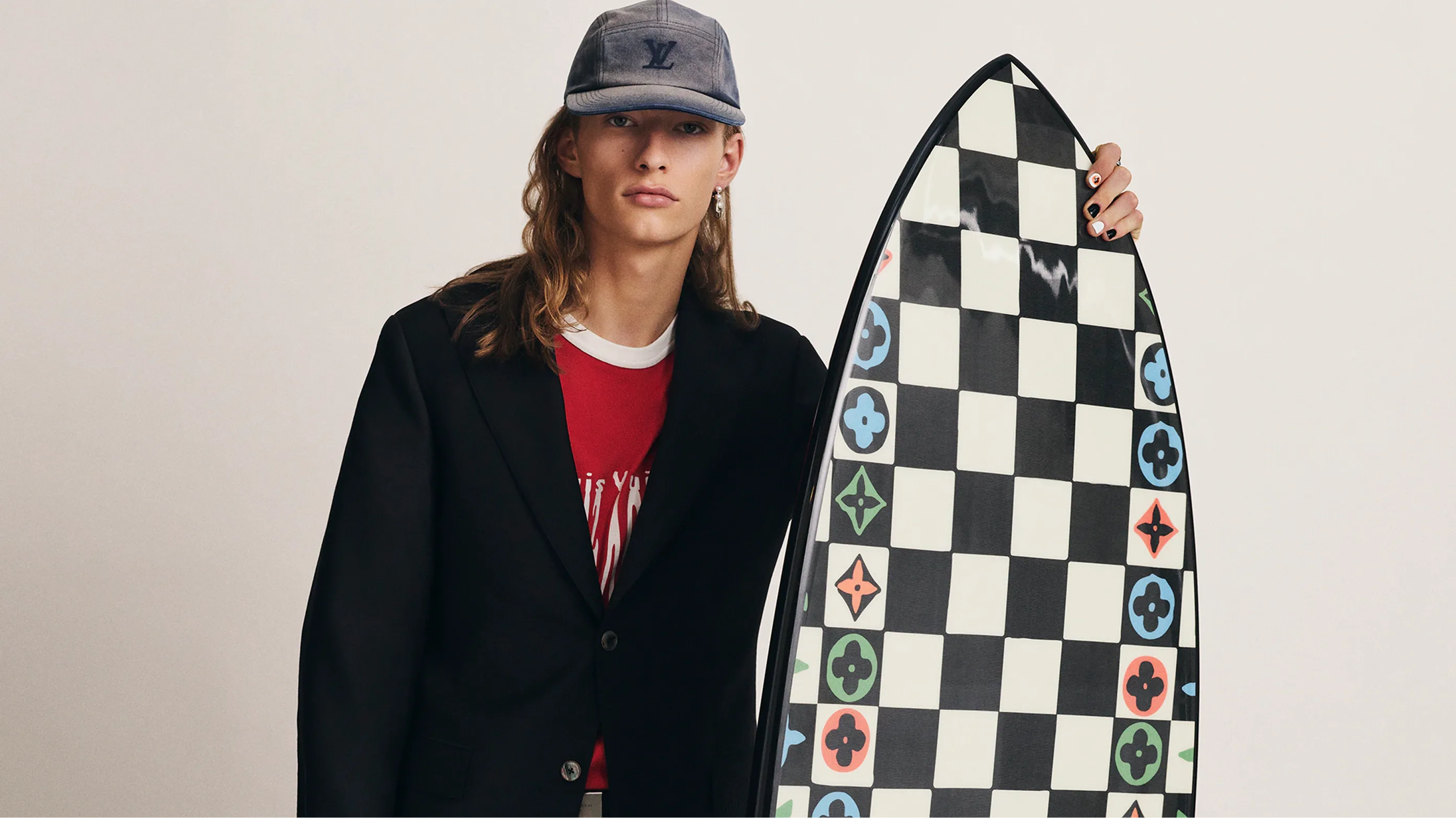 A person with long hair wearing a black blazer, red shirt, and baseball cap holds a black and white checkerboard surfboard decorated with colorful floral logos.