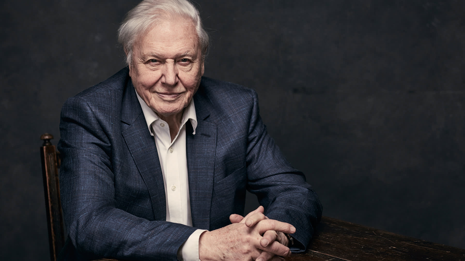 An elderly man with white hair sits at a dark wooden table, wearing a blue blazer over a white collared shirt, with his hands clasped and a slight smile, against a dark, plain background.