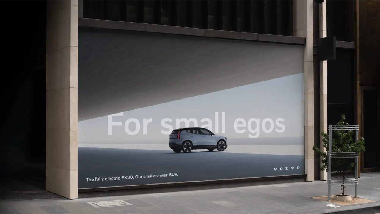 A Volvo billboard displays a compact silver SUV beneath the slogan For small egos with text reading, The fully electric EX30. Our smallest ever SUV. The advertisement is placed on the side of a city building.