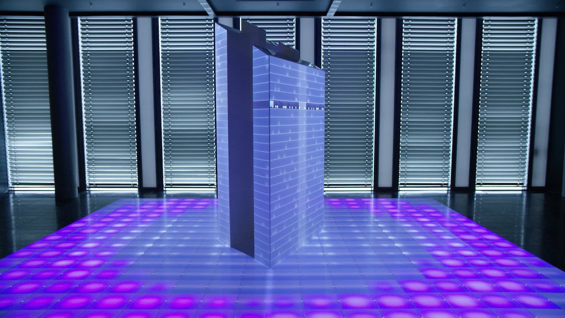 A tall, modern, rectangular server or electronic device stands on a glowing floor with purple and blue lights, in front of large windows with horizontal blinds in a sleek, futuristic room.