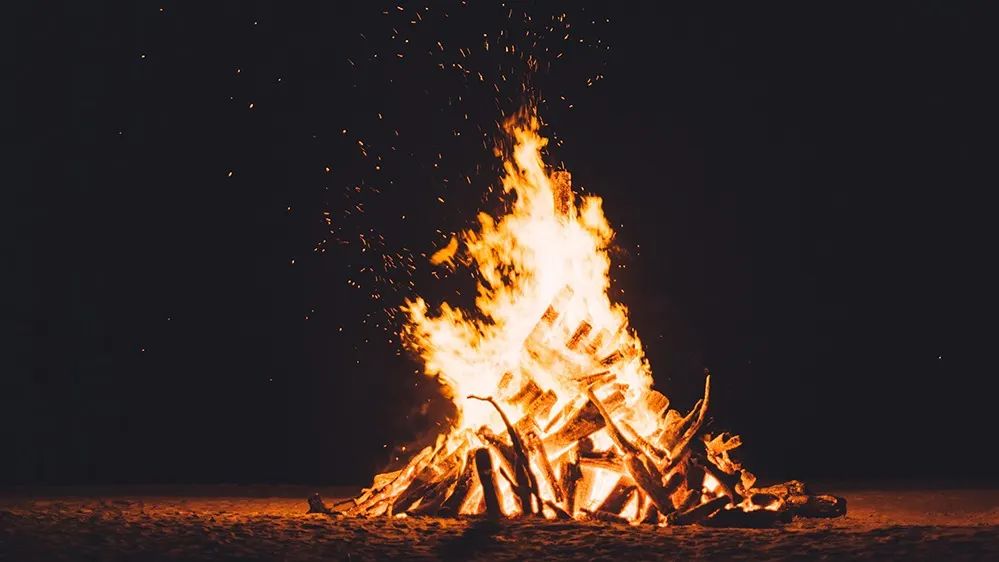 A large bonfire burns brightly at night, with flames rising from stacked wooden logs and glowing embers floating upward against a dark background.