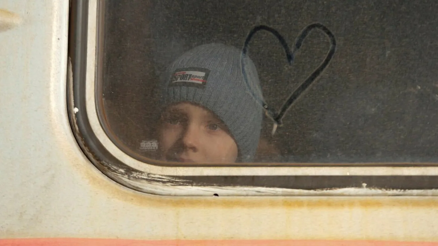 A child wearing a gray beanie looks out from behind a train window, with a heart shape drawn in the condensation on the glass.