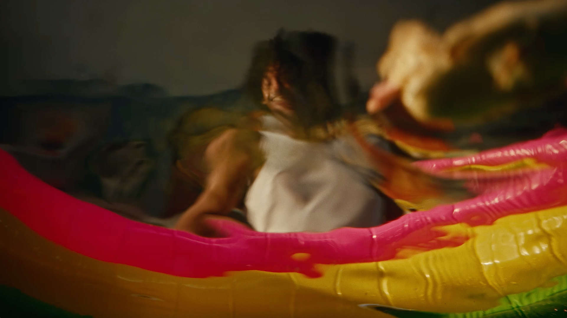A blurred, distorted image of a person in a white shirt seen through colorful, wavy plastic or glass, with prominent pink, yellow, and green sections in the foreground.