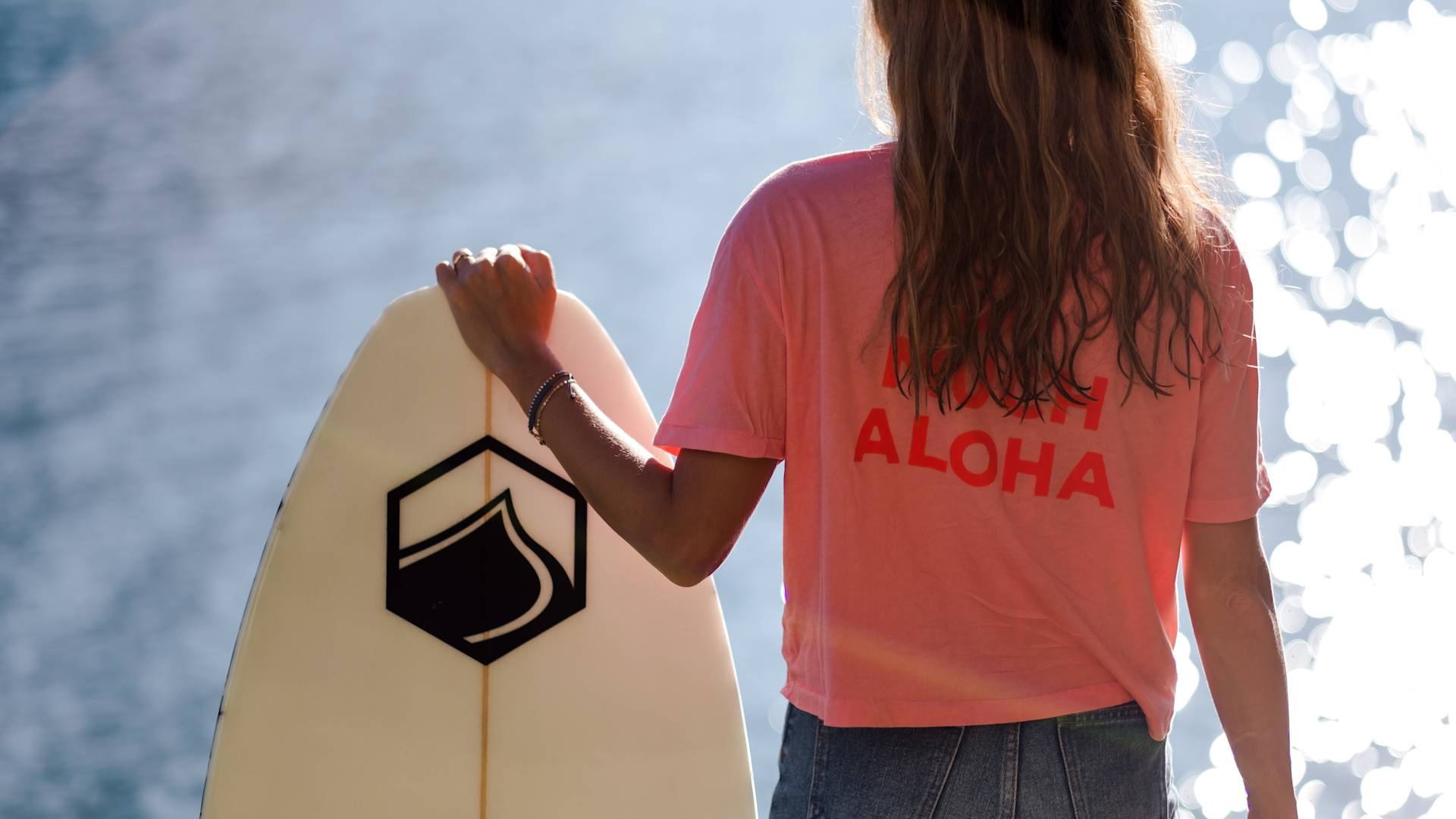 A person with long hair stands by the water, holding a surfboard. They wear a pink shirt with the word ALOHA printed on the back. The sunlight reflects off the water, creating a bright, summery scene.