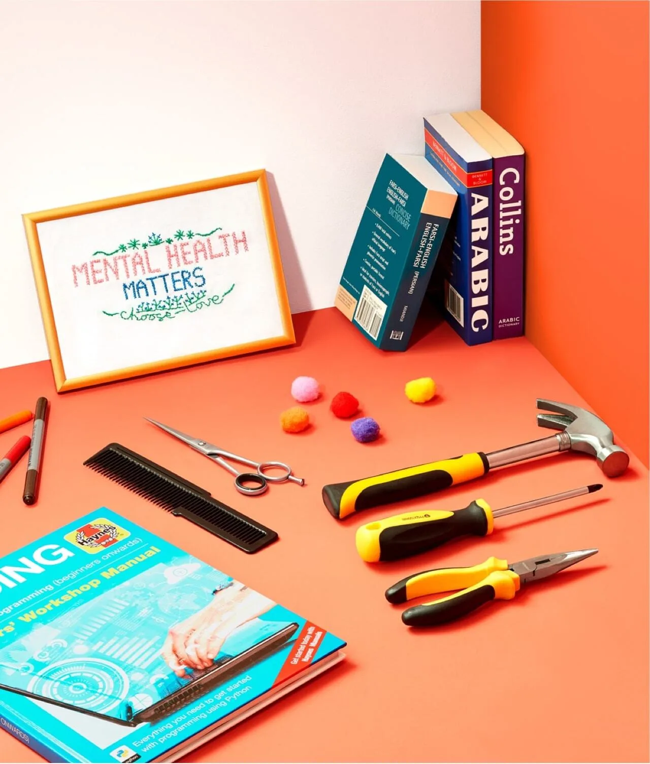 A desk with tools, pens, scissors, a comb, pom-poms, language dictionaries, a magazine, and a framed cross-stitch reading Mental Health Matters against an orange and white background.