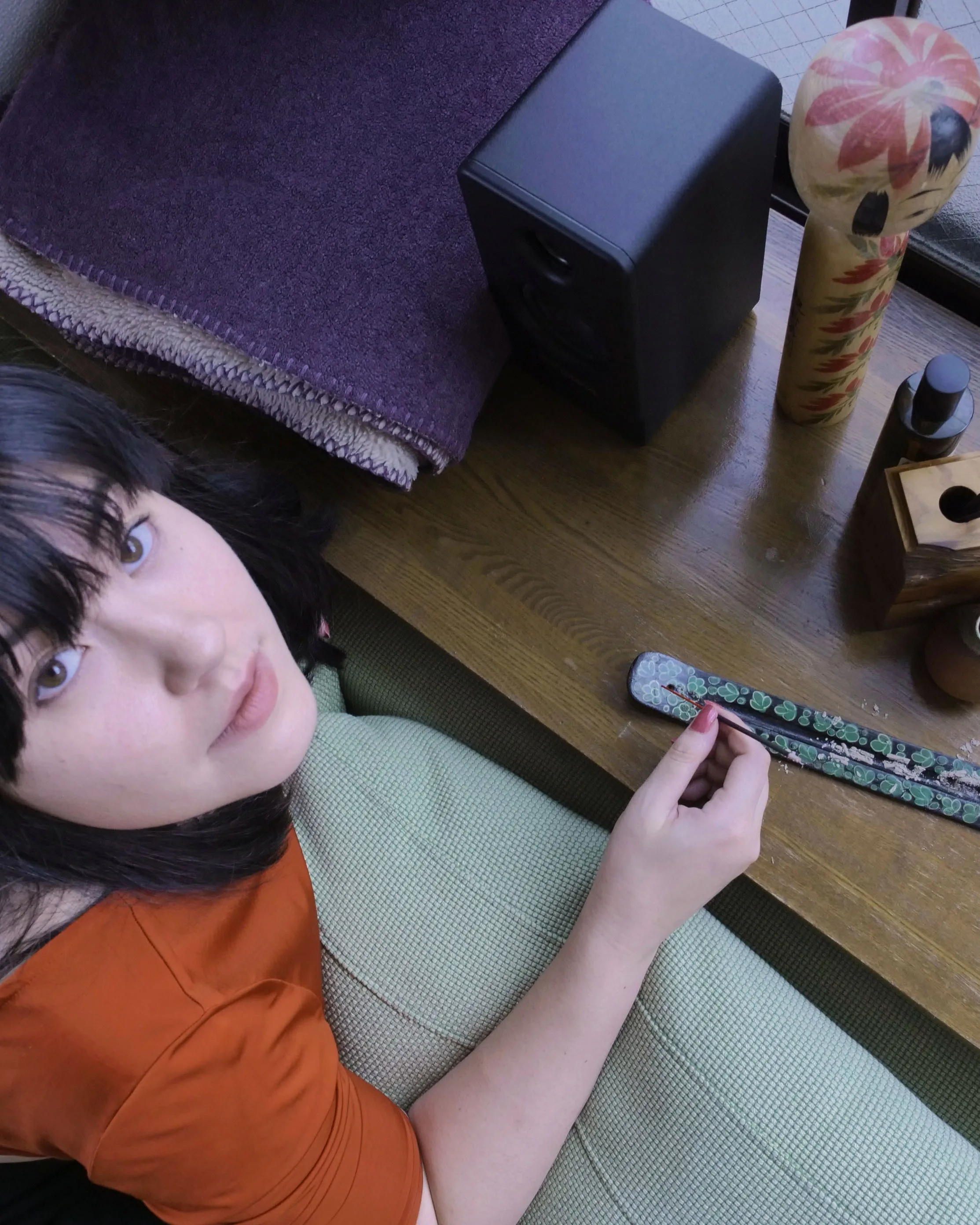 A person with dark hair and bangs, wearing an orange top, sits on a couch and looks up. On the wooden table beside them are a purple blanket, a speaker, a wooden doll, and decorative objects.