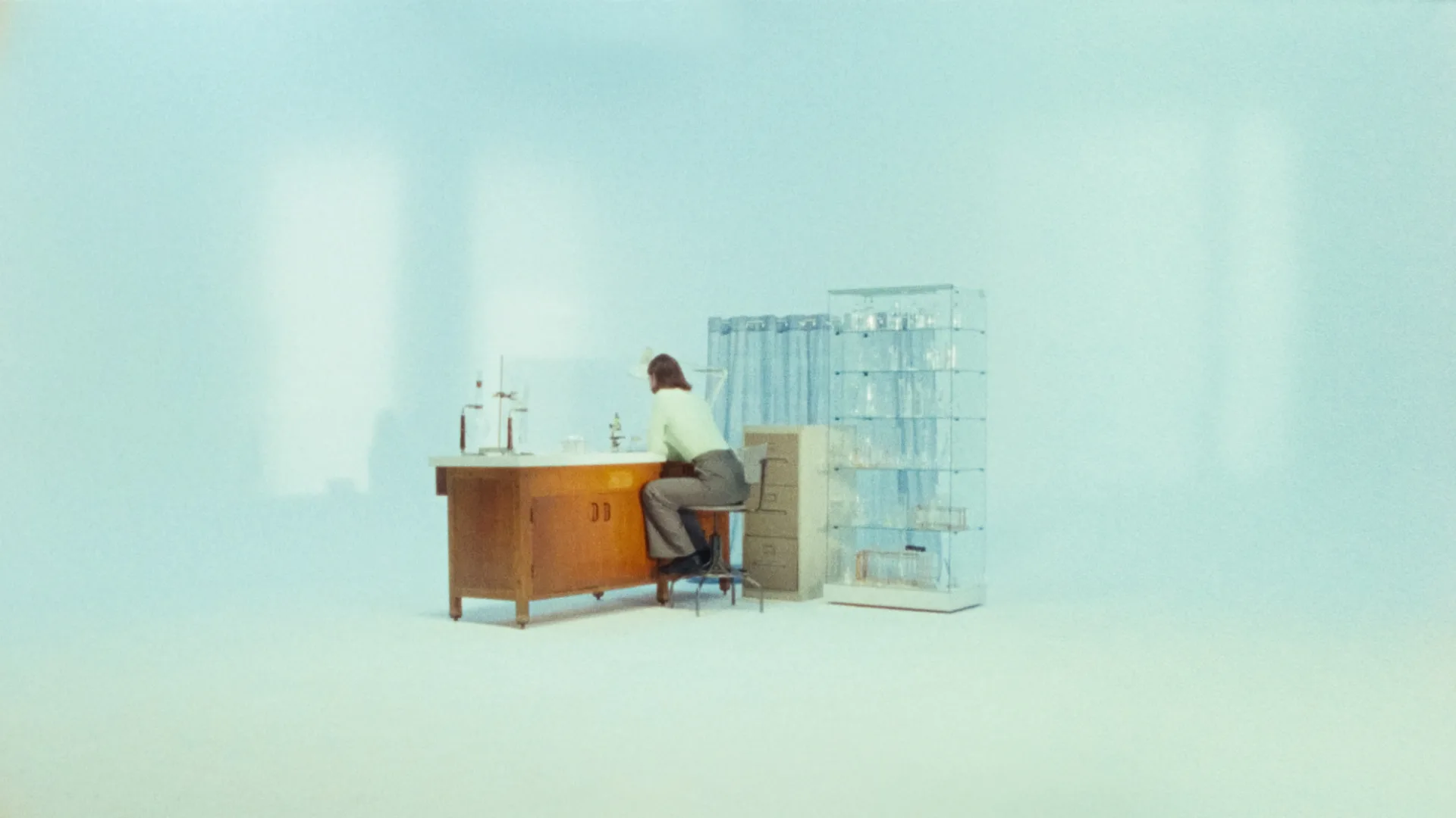 A person sits on a stool at a wooden desk in a minimalist, light-filled room with pale blue walls. Laboratory glassware and filing cabinets are arranged nearby, creating a sparse, scientific atmosphere.