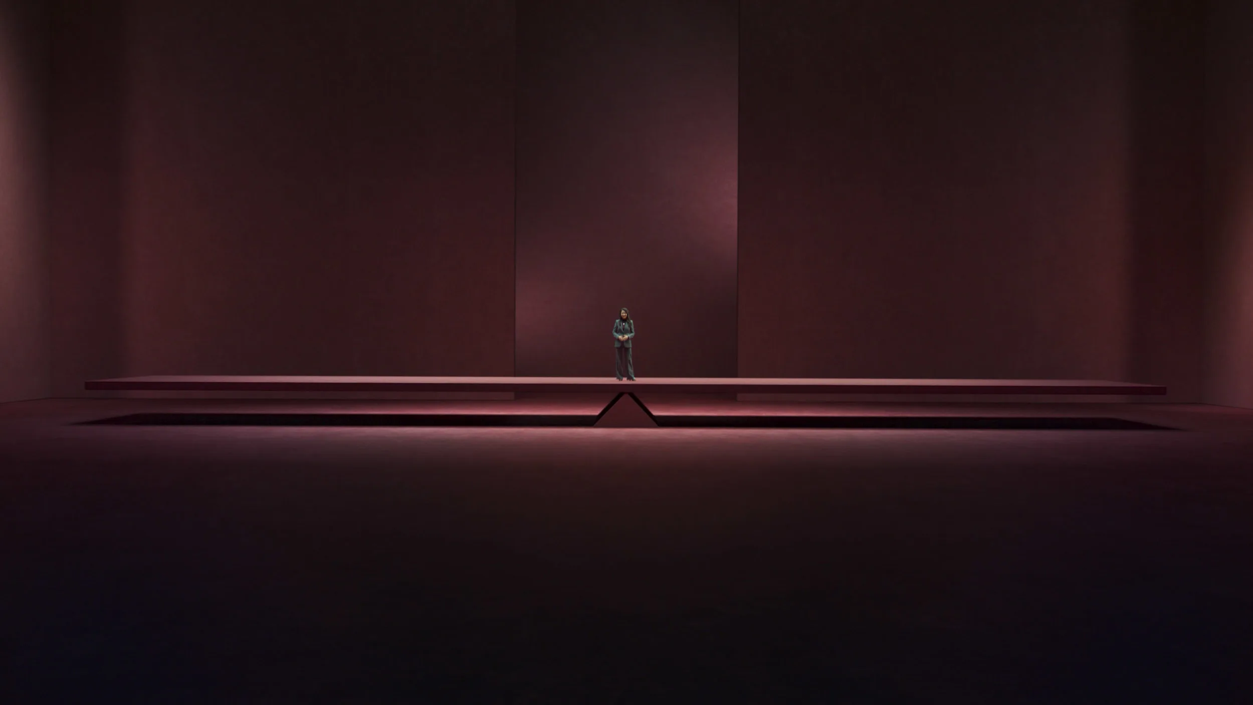 A person stands alone on a large, minimalist stage with deep red walls and dramatic lighting, emphasizing the emptiness and scale of the space around them.