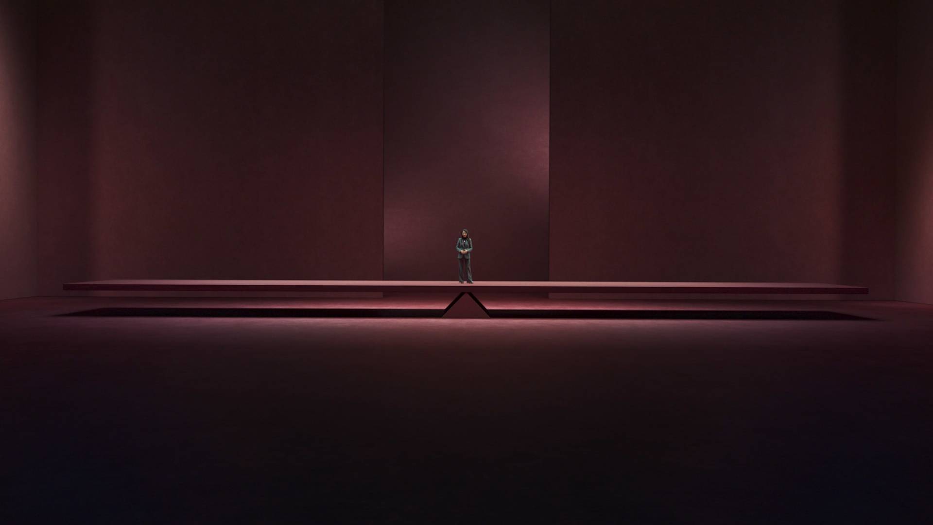 A person stands alone on a large, minimalist stage with deep red walls and dramatic lighting, emphasizing the emptiness and scale of the space around them.
