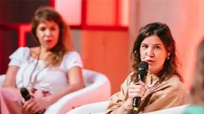 Two women are seated in white chairs, engaged in a discussion. The woman in the foreground is holding a microphone and speaking, while the woman in the background listens. The setting has warm, red-toned lighting.