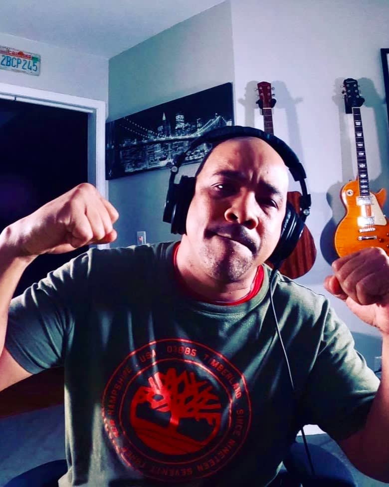 A man wearing headphones and a green Timberland t-shirt flexes his arms and makes a serious face. Behind him are two guitars mounted on the wall and a black-and-white cityscape photo.