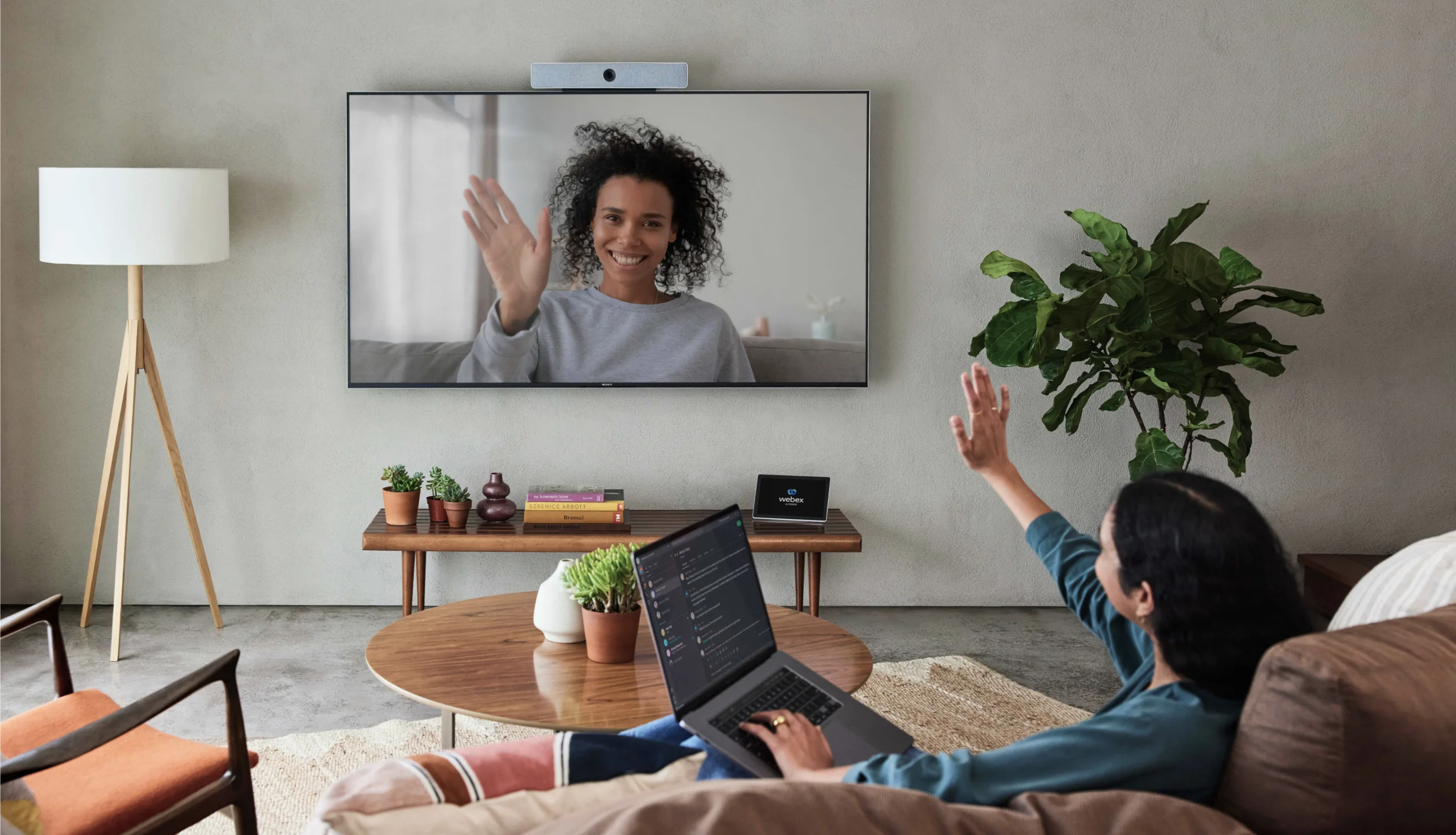 A person sits on a couch with a laptop, waving at a woman on a large wall-mounted screen during a video call. The living room has plants, a coffee table, and modern decor.