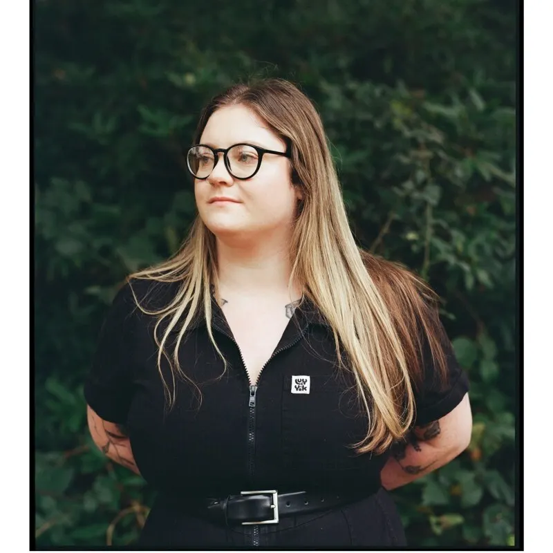 A woman with long, blonde hair and glasses stands in front of green foliage, wearing a black zip-up top and a belt, looking slightly to the side with her hands behind her back.