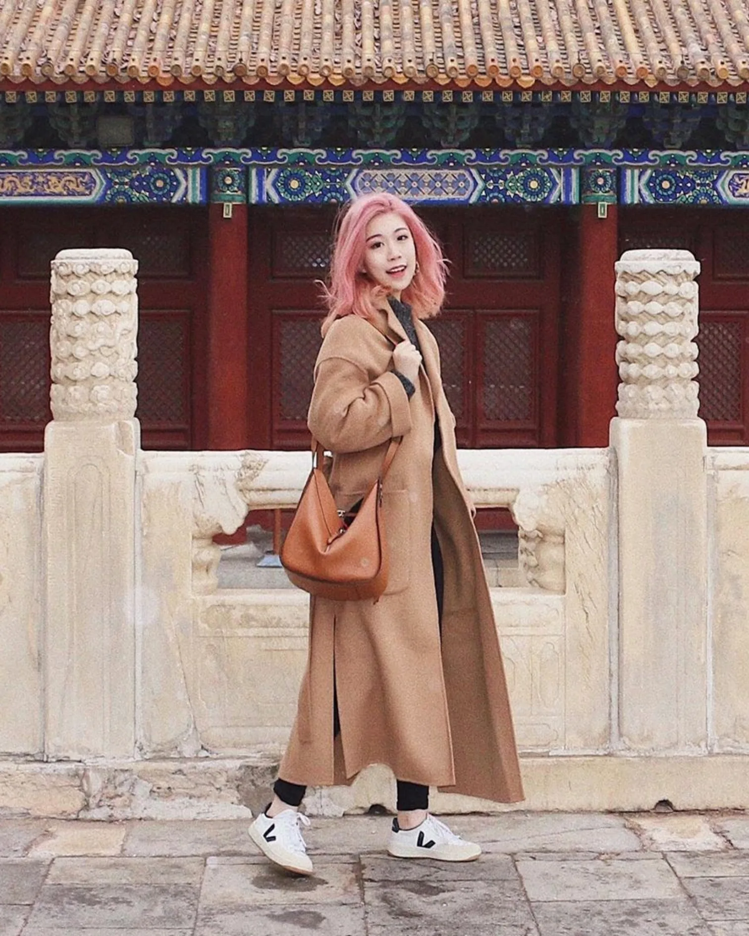 A woman with pink hair, wearing a long tan coat, black pants, white sneakers, and carrying a brown bag, poses in front of an ornate building with traditional Asian architectural details and red doors.
