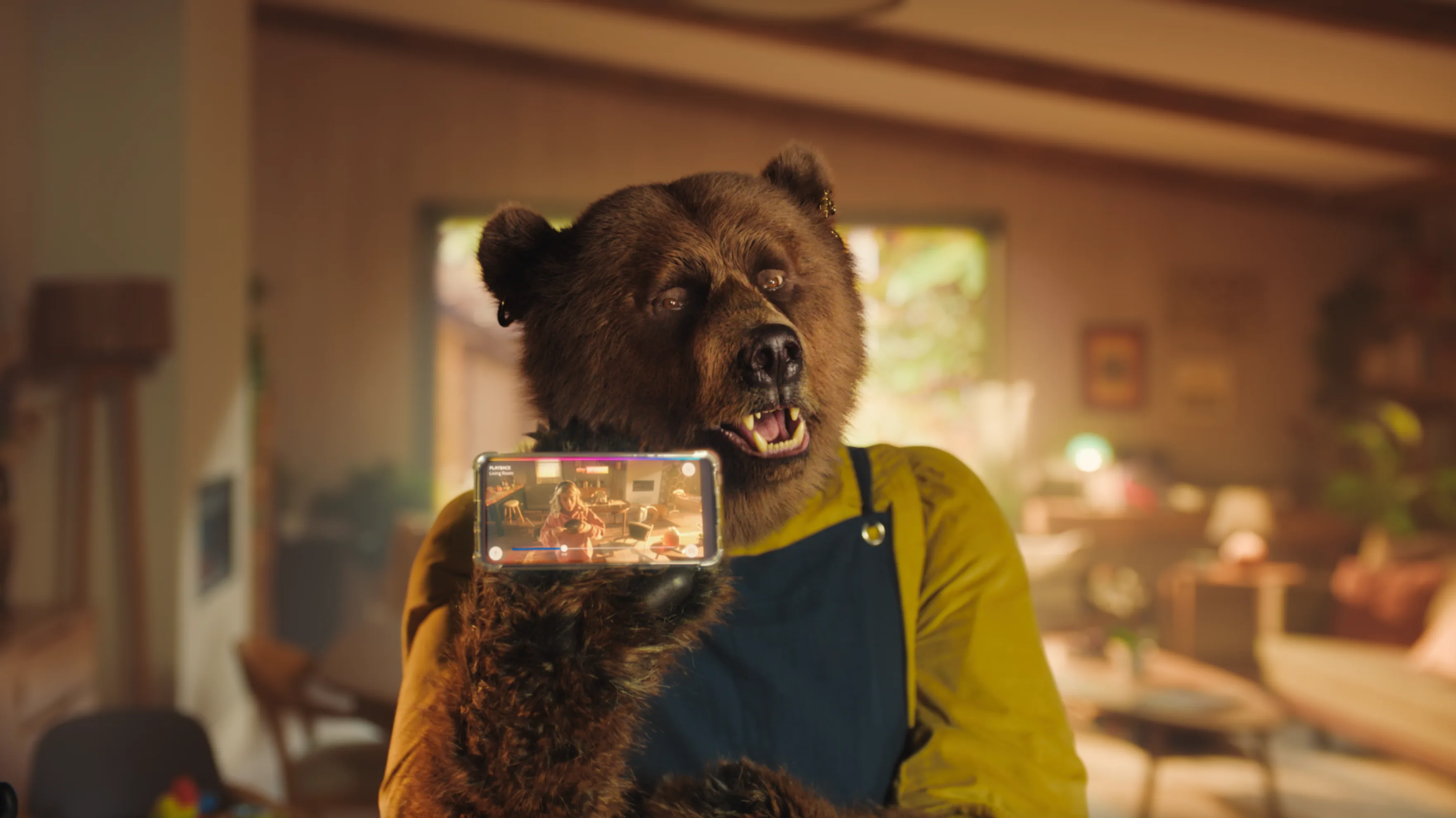 A bear wearing a yellow shirt and blue apron holds a smartphone, showing its own image on the screen, inside a cozy, sunlit living room.